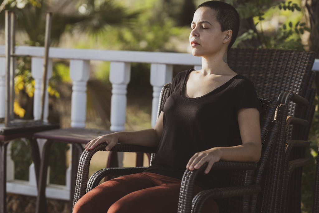 A woman in meditation with eyes closed sitting on a chair outdoors in the sunlight.